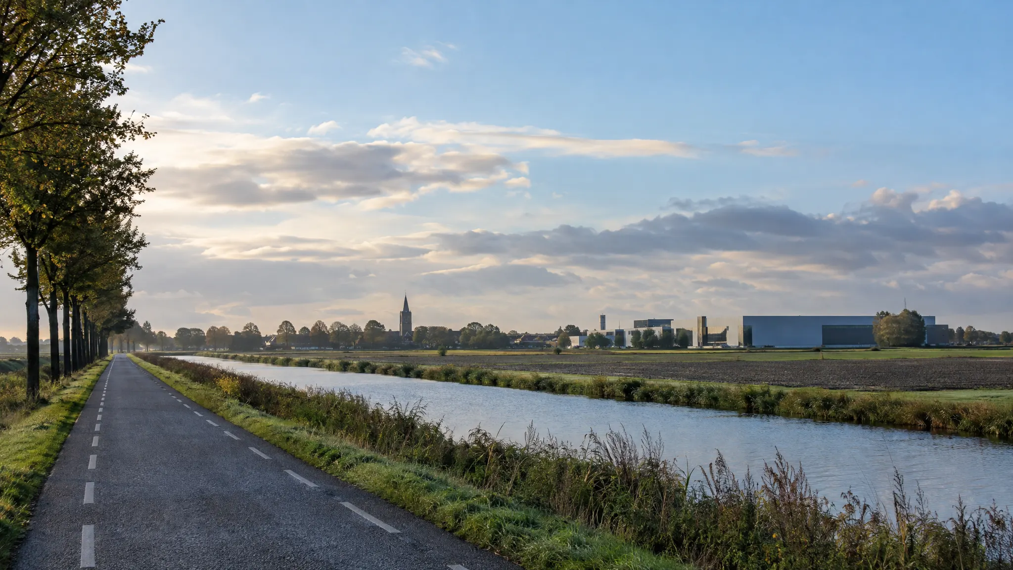 Het vlakke Groninger land met een kanaal, een bomenrij en in de verte een kerktoren en een bedrijfspand.